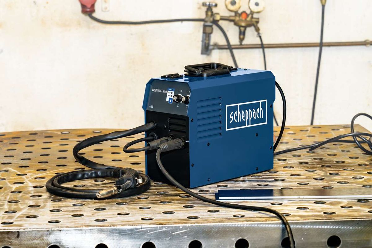Scheppach Multischweißgerät WSE4000-Multi auf perforiertem Metalltisch. Kabel und Schlauch liegen auf dem Tisch. Im Hintergrund sind Gasflaschen und Schläuche zu sehen.