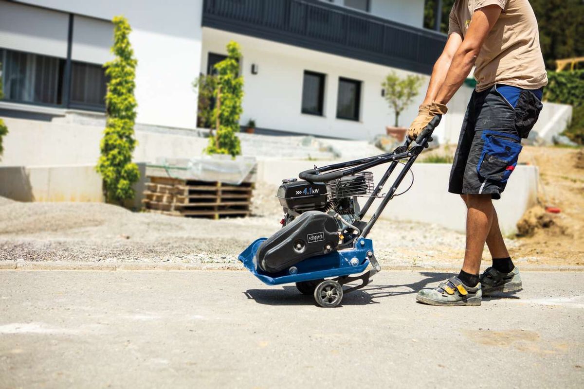 Ein Mann bedient eine Scheppach Rüttelplatte HP1300S auf einer Betonfläche. Die Maschine hat einen schwarzen Motor und ein blaues Gehäuse. Im Hintergrund sind ein Gebäude und Paletten zu sehen.