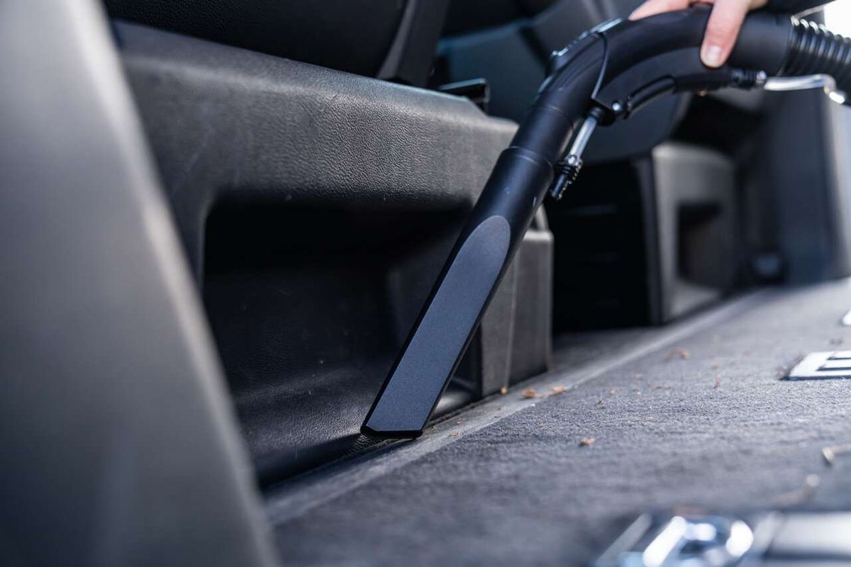 Ein Scheppach SprayVac20-Sauger, der in den Kofferraum eines Autos gehalten wird. Der Sauger hat einen schwarzen Griff und einen grauen Behälter. Der Sauger wird von einer Hand gehalten, die den Griff festhält.