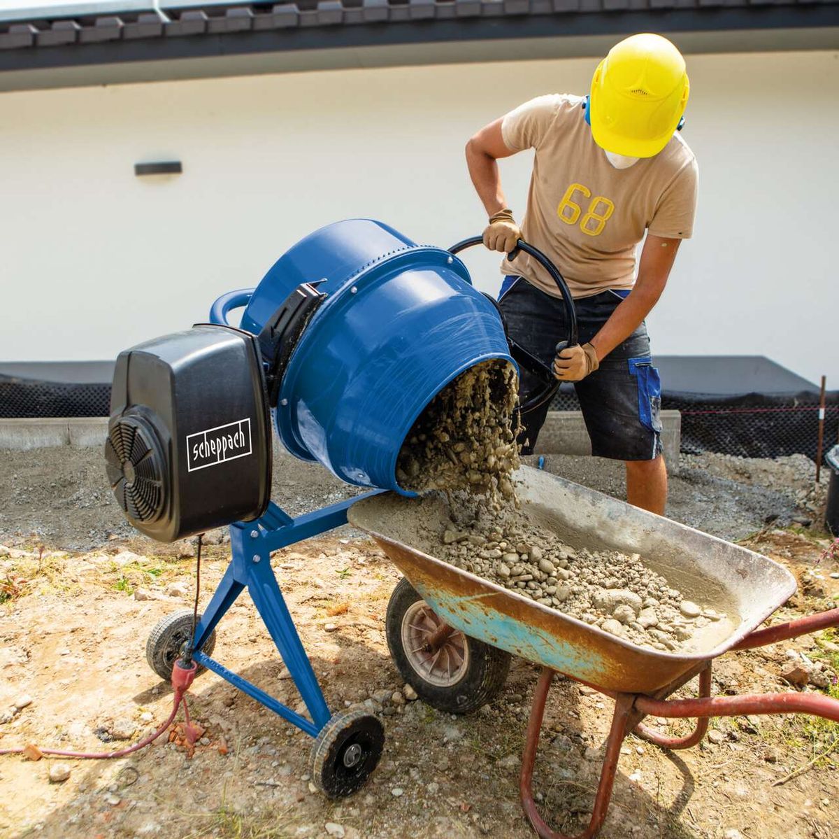 Ein Arbeiter mit gelbem Helm bedient einen Betonmischer von Scheppach. Der Mischer füllt eine Schubkarre mit Beton. Der Mischer hat ein blaues Gehäuse und einen schwarzen Motor mit dem Scheppach-Logo. Die Szene spielt auf einer Baustelle.