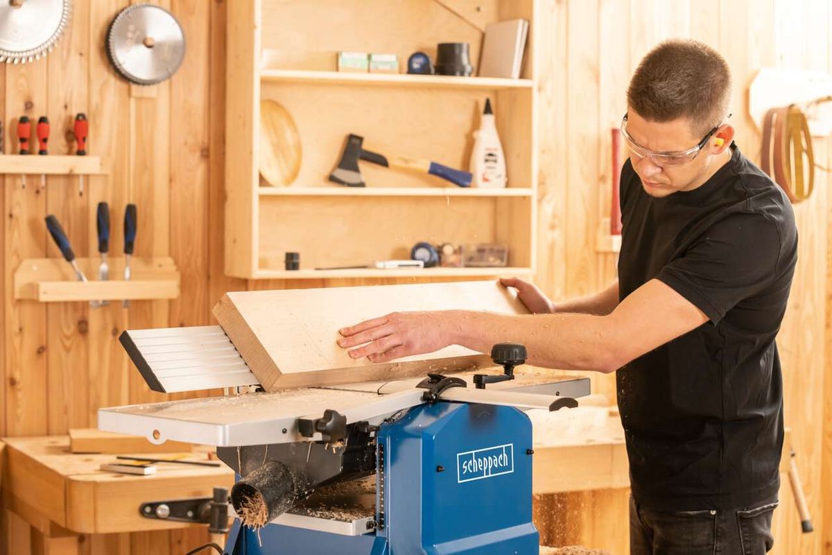 Ein Mann bedient die Abricht- & Dickenhobelmaschine HMS1070 von Scheppach. Er trägt Schutzbrille und bearbeitet ein Holzbrett. Im Hintergrund sind Werkzeuge und Holzregale zu sehen.