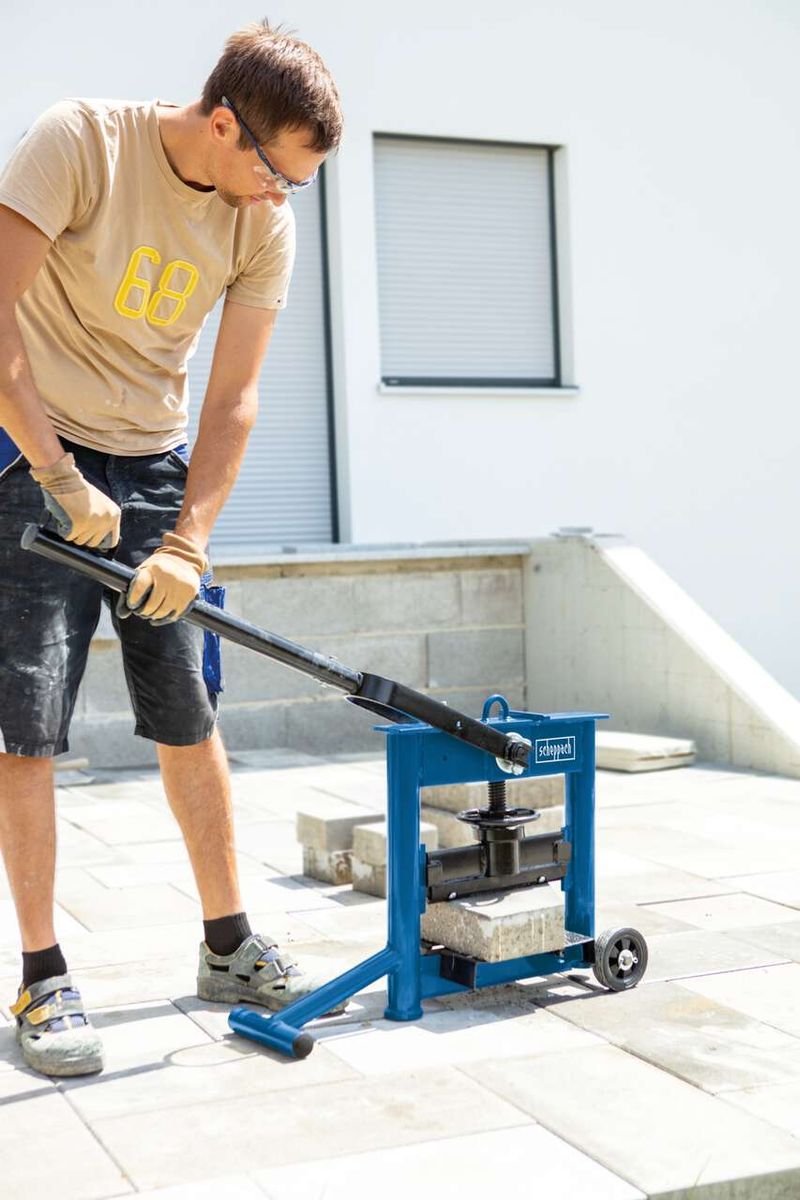 Ein Mann bedient einen Steinknacker HSC130 von Scheppach. Er trägt Schutzhandschuhe und -brille und drückt den Hebel des Geräts nach unten. Der Steinknacker steht auf einer Betonplatte.