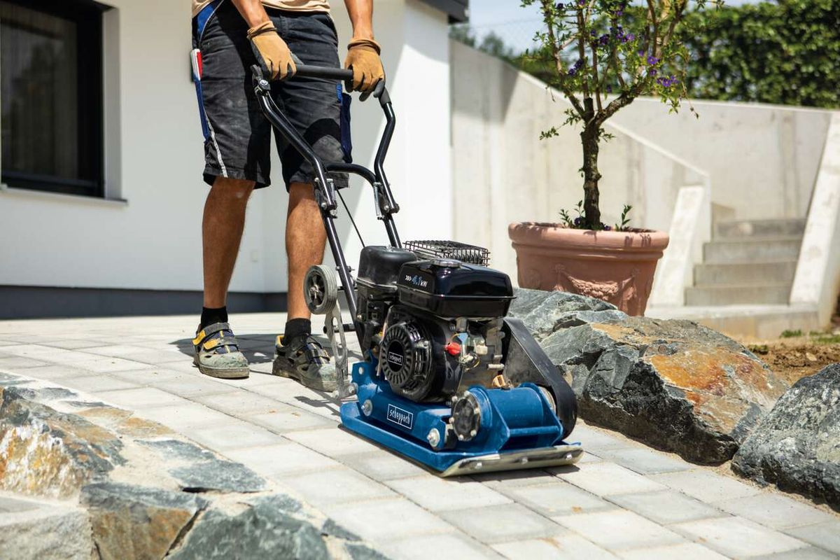 Ein Mann bedient eine Scheppach Rüttelplatte HP1300S auf einem gepflasterten Boden. Die Maschine hat einen schwarzen Motor und eine blaue Basis. Im Hintergrund sind ein Blumentopf und eine Hauswand zu sehen.