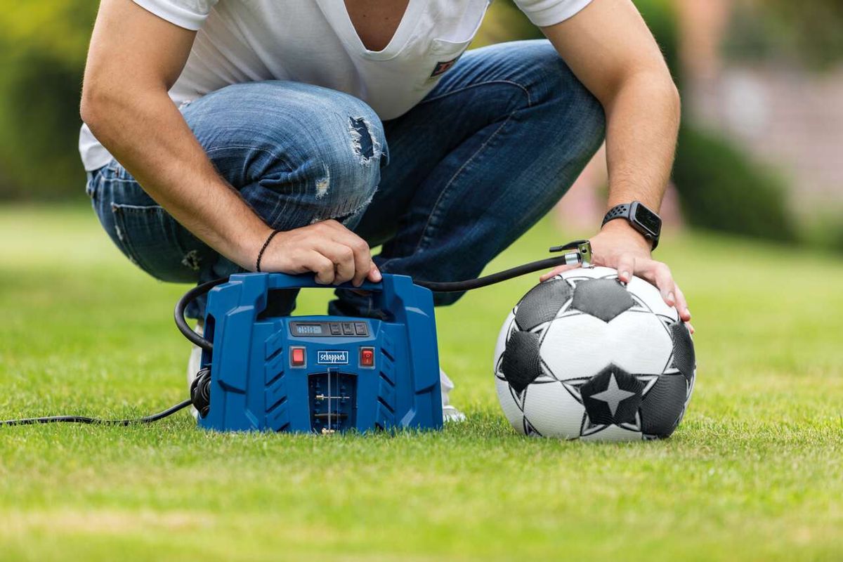 Ein Mann sitzt auf einem Rasen und verwendet einen Scheppach-Kompressor, um einen Fußball aufzublasen. Der Kompressor ist blau und hat ein digitales Druckdisplay.