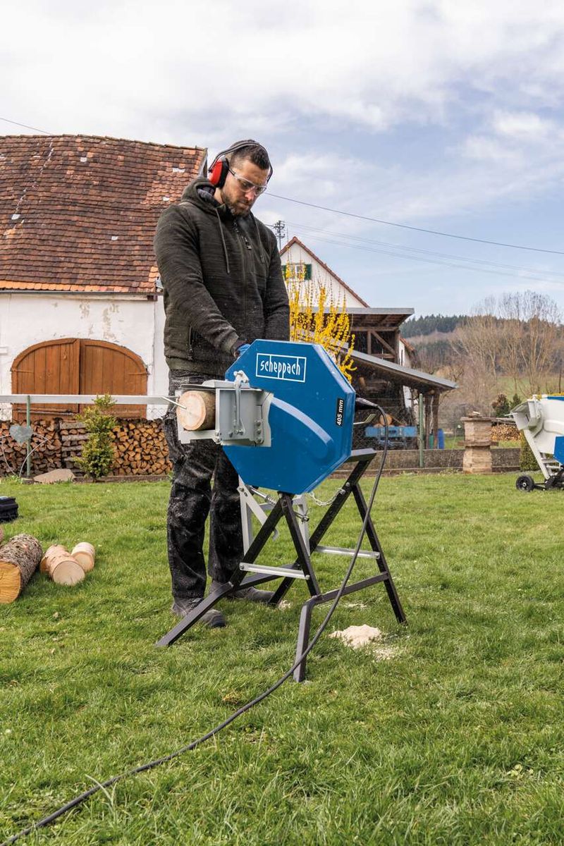 Ein Mann bedient eine Wippsäge HS410 von Scheppach im Freien. Die Säge ist auf einem Ständer montiert und wird zum Schneiden von Holzstämmen verwendet. Im Hintergrund sind ein Schuppen und weitere Werkzeuge zu sehen.