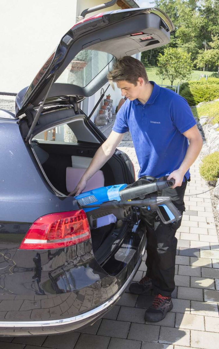 Ein Techniker platziert ein Zubehörteil in den Kofferraum eines Autos. Er trägt ein blaues Hemd mit dem Aufdruck 'Scheppach'. Der Hochdruckreiniger HCE2200 von Scheppach liegt im Kofferraum.