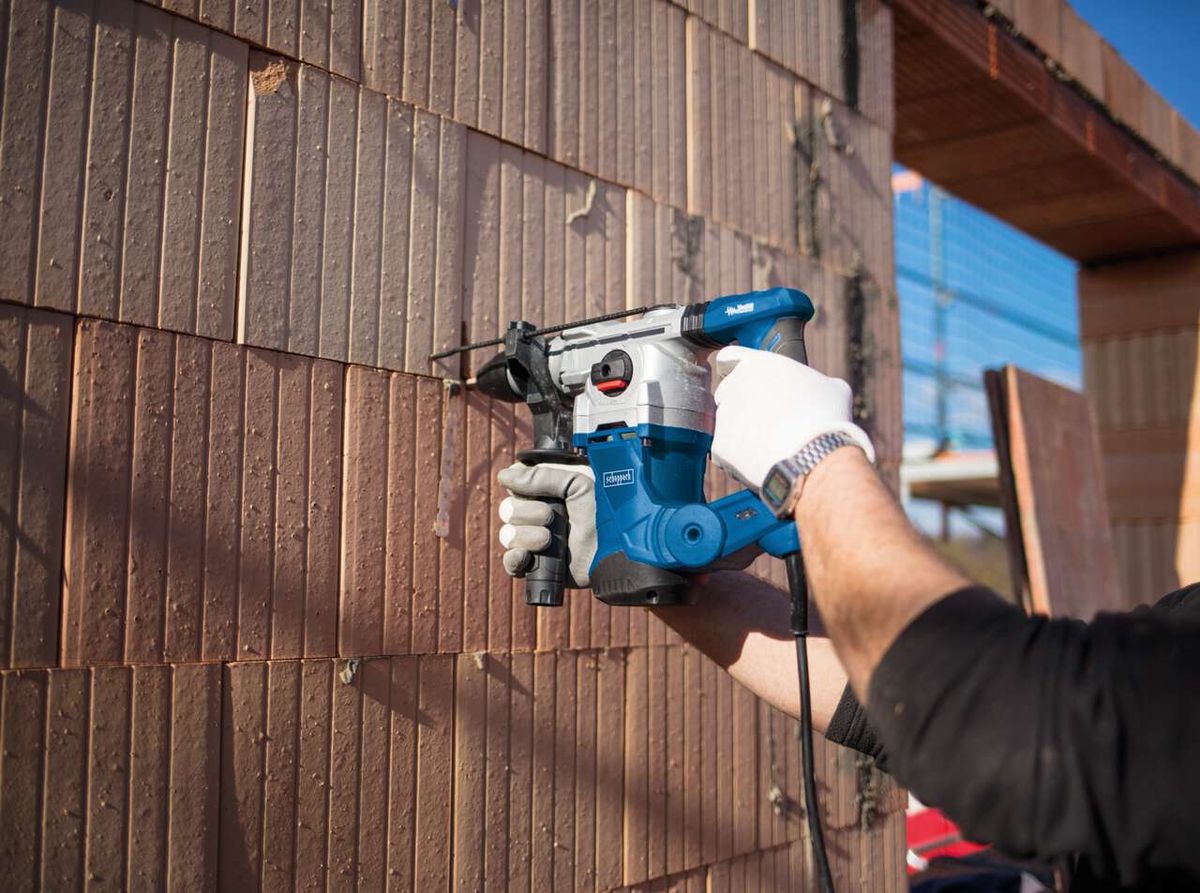 Bohrhammer DH1300 Plus von Scheppach im Einsatz. Ein Arbeiter hält das Werkzeug und bohrt in eine Wand aus Holzpaneelen. Der Bohrhammer ist Teil eines DIY-Sets mit Koffer und Zubehör.