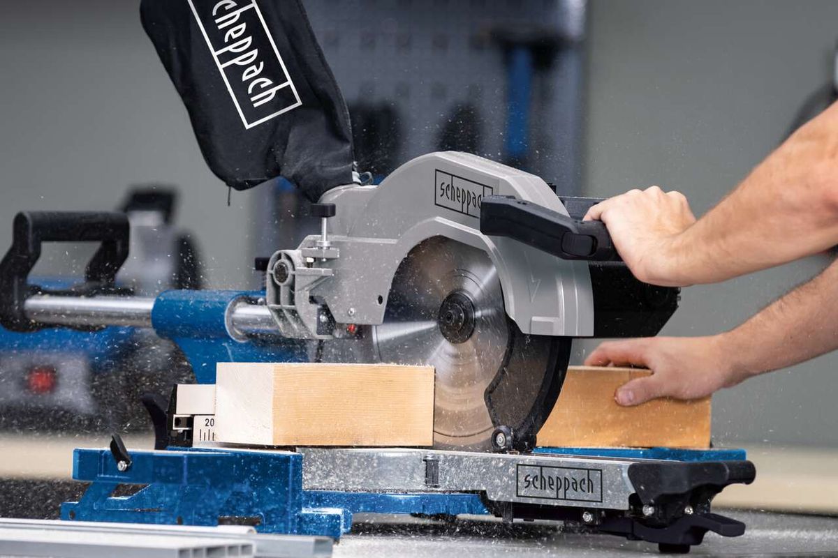 Kappzugsäge HM110MP von Scheppach im Einsatz. Ein Benutzer schneidet ein Holzbrett. Die Säge ist auf einem blauen und schwarzen Tisch montiert.