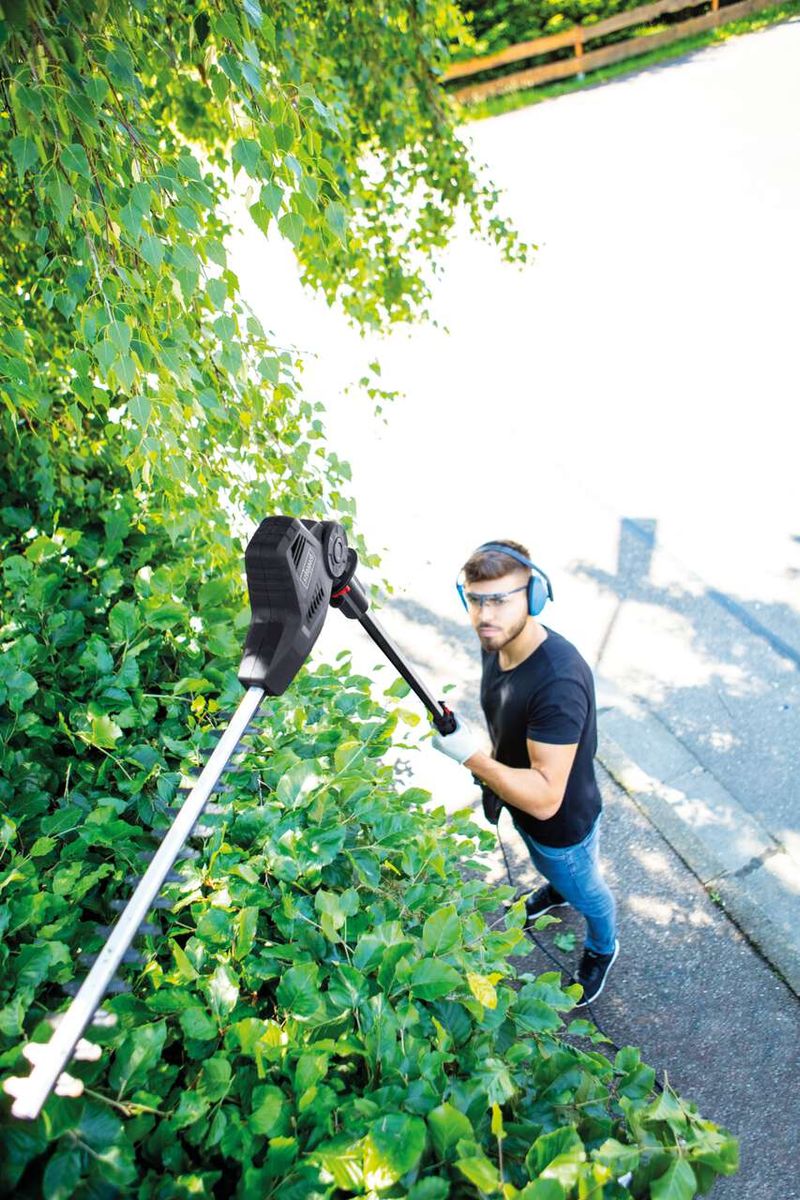 Ein Mann schneidet mit einer Elektro Heckenschere TPX710 von Scheppach einen grünen Strauch. Er trägt Schutzbrille und Kopfhörer.