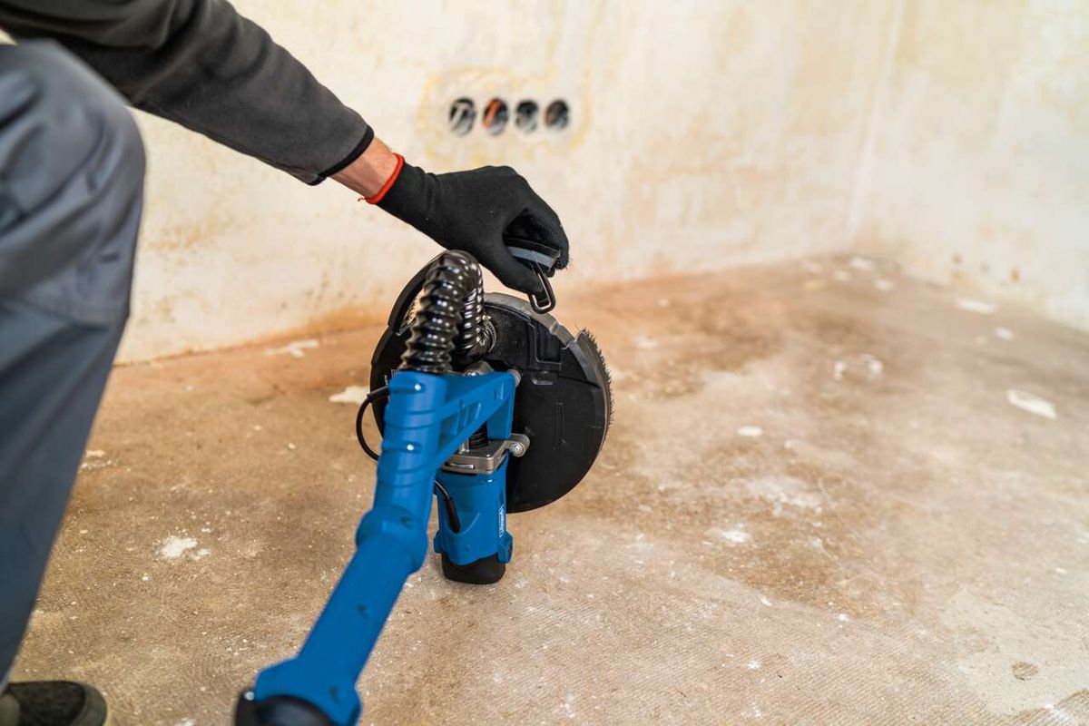 Wand-Deckenschleifer DS920X von Scheppach im Einsatz. Person hält das Gerät mit Handschuhen und führt es über den Boden. Der Schleifer hat einen blauen Griff und eine schwarze Schleifscheibe.