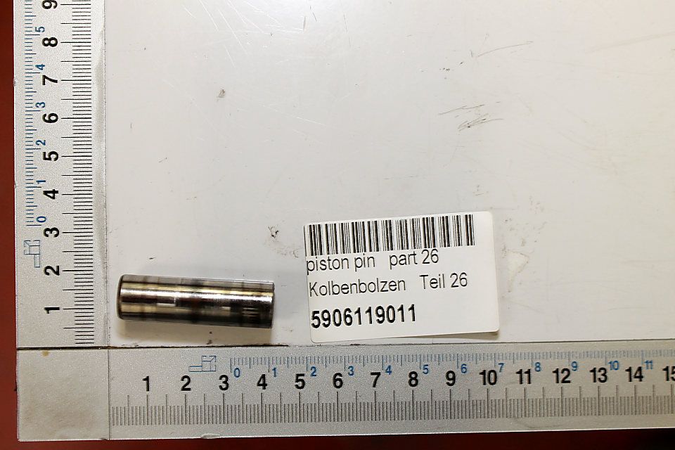 Ein Kolbenbolzen mit der Bezeichnung 'Kolbenbolzen Teil 26' und der Artikelnummer 5906119011. Der Bolzen liegt neben einem Maßstab und einem Beschriftungslabel. Der Kolbenbolzen ist von der Seite zu sehen.