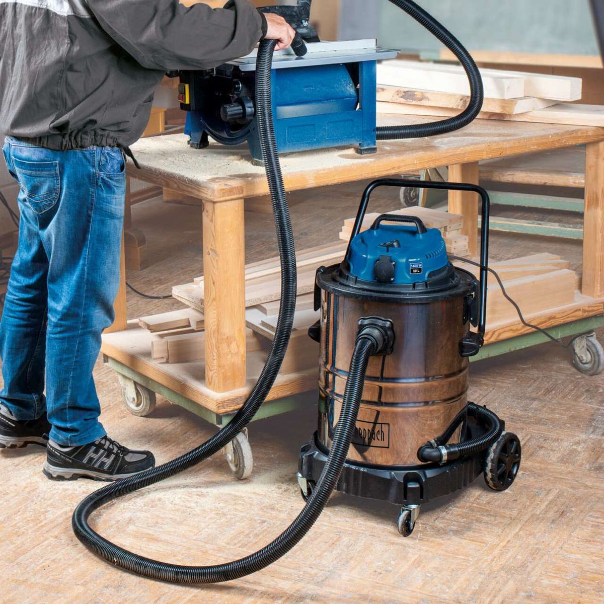 Der Scheppach Nass- und Trockensauger ASP50-ES mit 50L, HEPA-Filter, Steckdose und Einschaltautomatik. Der Sauger ist mit einem Holzfäller verbunden, um Sägespäne zu entfernen.