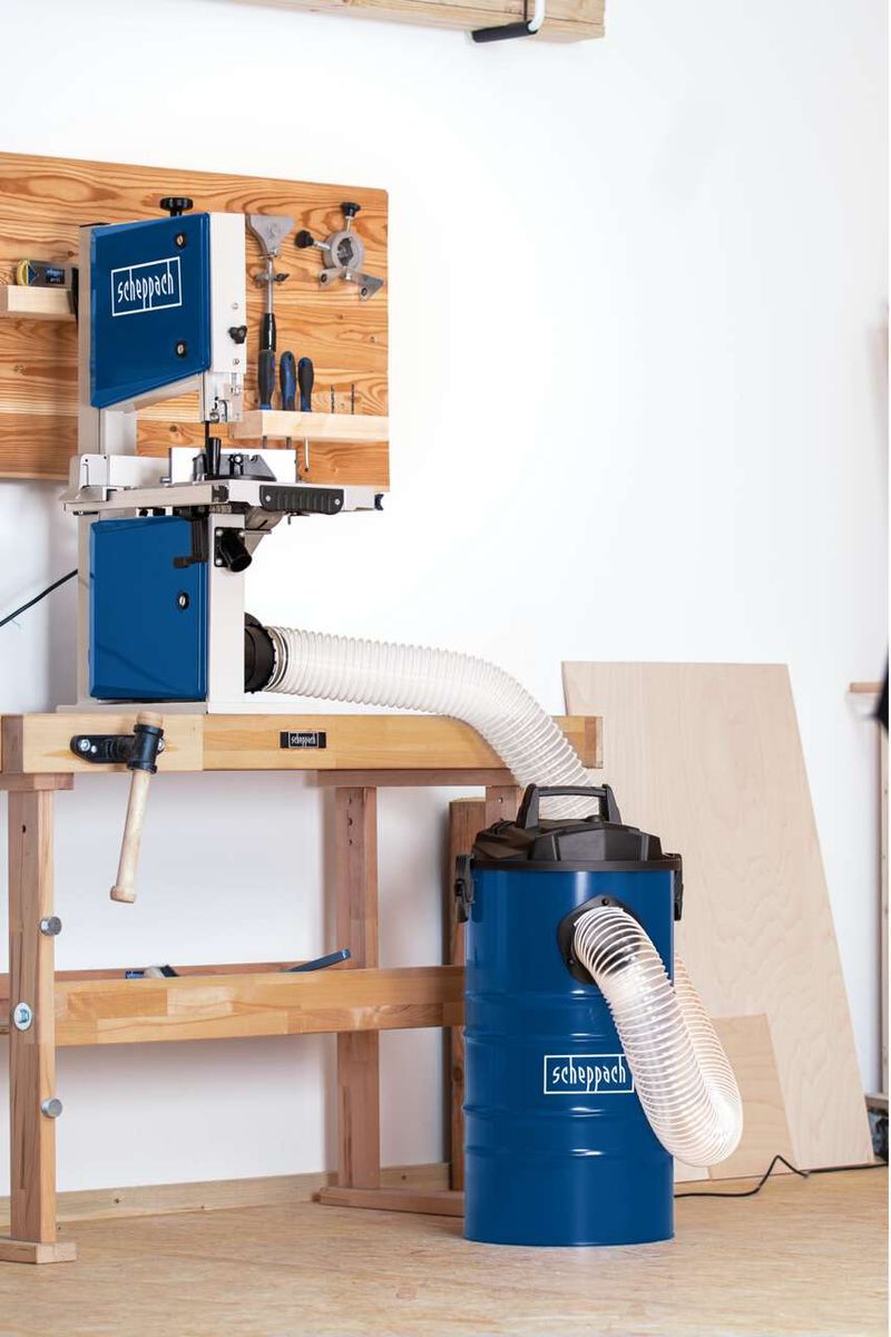 Scheppach DC100 dust extractor with vacuum and hose, mounted on a workbench with wooden shelves and tools in the background.
