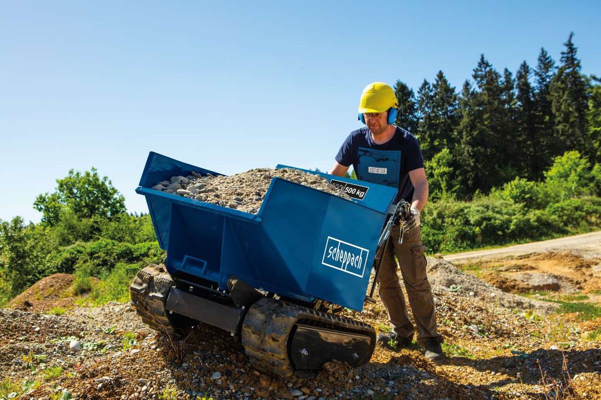 Kettendumper DP5000 Scheppach mit 500 kg Traglast. Ein Arbeiter in Schutzausrüstung steht neben dem beladenen Dumper auf einem Baustellengelände. Im Hintergrund sind Bäume und eine unbefestigte Straße zu sehen.