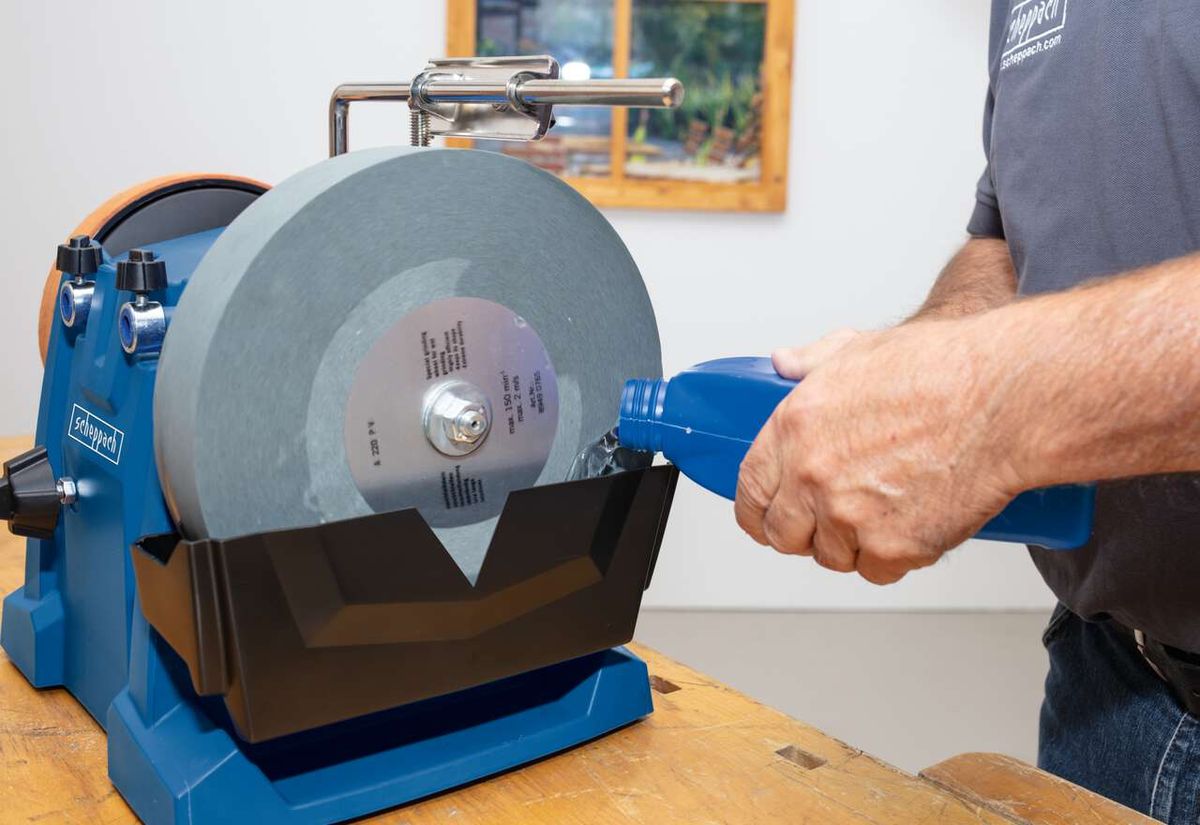 Scheppach Wet Bench Grinder Tiger 7000S in use. A user holds an object against the grinding wheel. The grinding wheel is 250mm in diameter with a grit of 220.