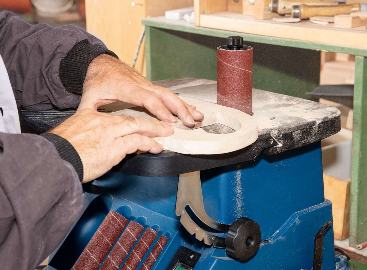 Ein Mann bedient einen Scheppach OSM600 Oszillierender Kanten-, Band- und Spindelschleifer. Er hält ein Werkstück gegen die rotierende Schleifscheibe.