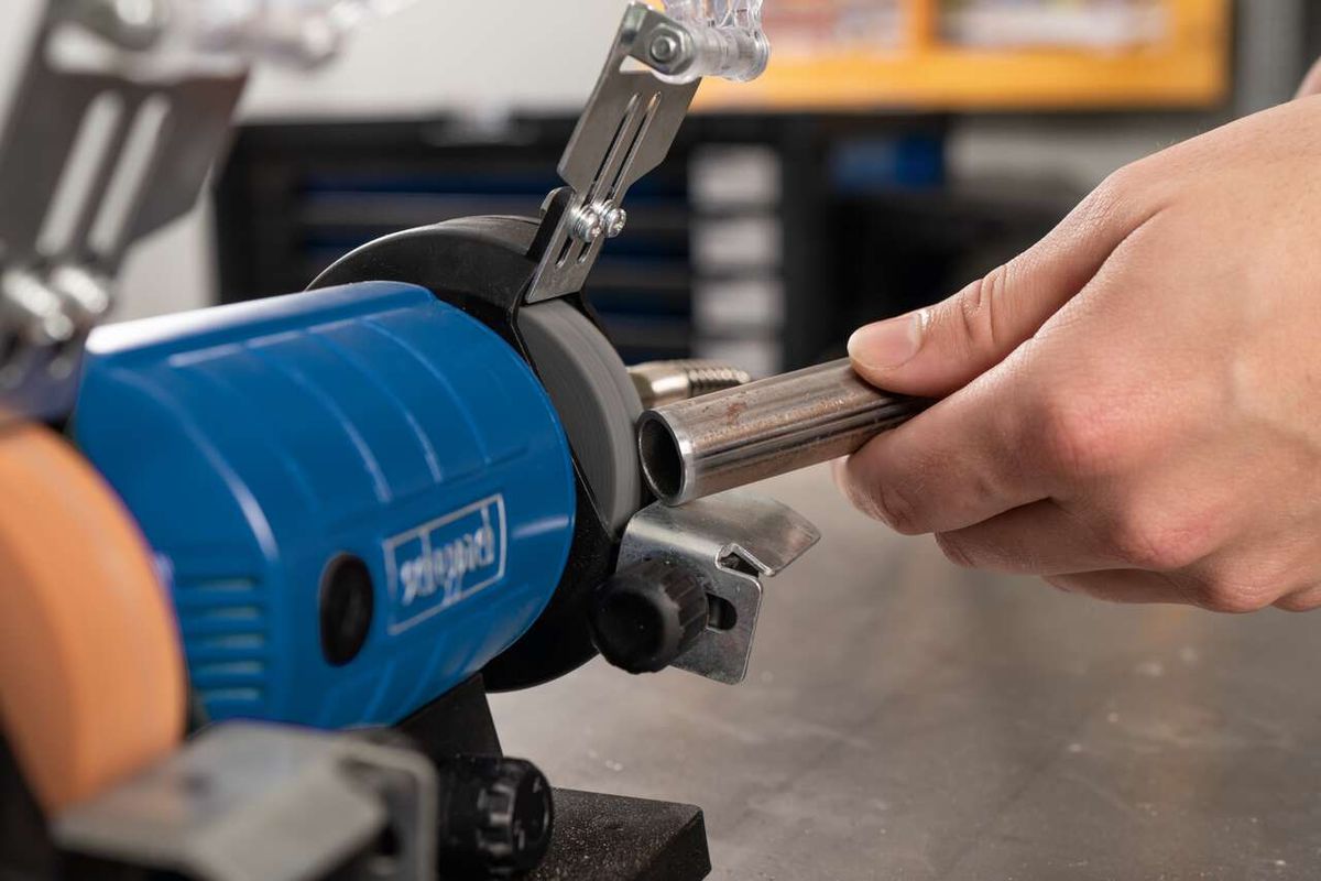 Ein Benutzer hält einen Metallstab in einen Doppelschleifer von Scheppach. Der Schleifer ist eingeschaltet und bearbeitet den Stab.