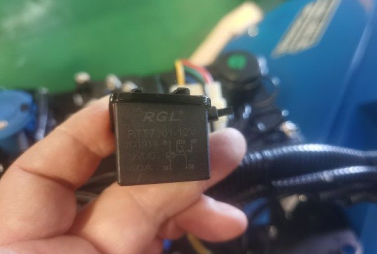 Ein Hand hält ein Relais für Kontrollpanel RTT7101-12V / JD1914 / 40A von Scheppach. Das Relais ist schwarz und hat Beschriftungen mit technischen Details. Im Hintergrund ist ein blaues Kontrollpanel zu sehen.
