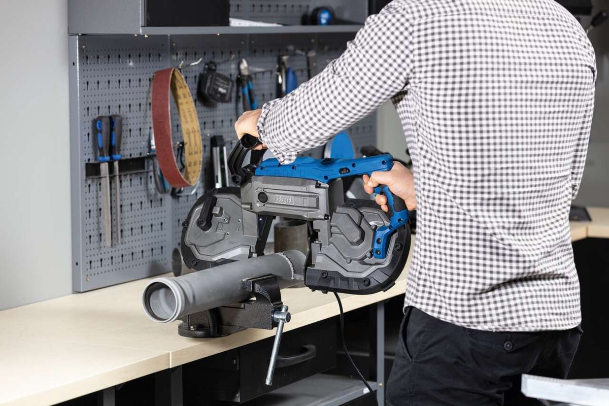 Metallbandsäge MBS1200xt von Scheppach, seitliche Ansicht. Ein Mann hält die Bandsäge fest, während sie auf einem Tisch montiert ist. Im Hintergrund sind Werkzeuge an der Wand befestigt.