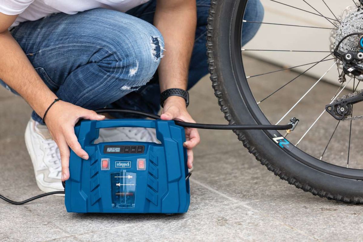 Ein Mann sitzt auf dem Boden und verwendet einen tragbaren Kompressor von Scheppach, um ein Fahrrad aufzubauen. Der Kompressor ist mit dem Fahrrad verbunden und zeigt 2,25 bar an.