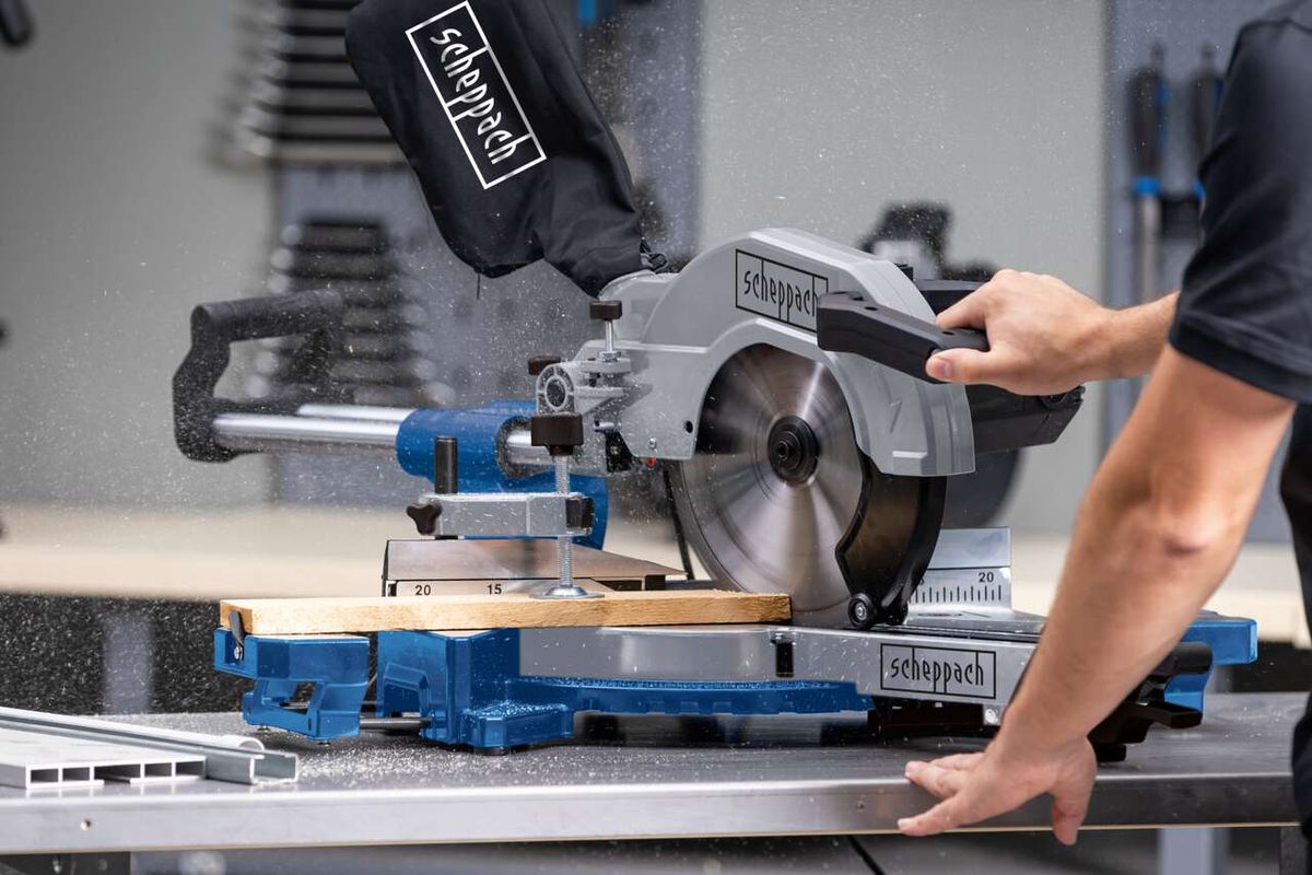 Kappzugsäge HM110MP von Scheppach im Einsatz. Ein Benutzer führt ein Holzstück durch die Säge. Die Säge ist auf einem blauen Tisch montiert und schneidet präzise.