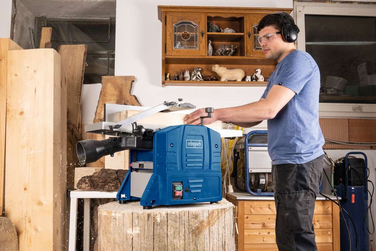 Ein Mann bedient eine Scheppach Abricht- und Dickenhobel HMS860 in einer Werkstatt. Die Maschine hat einen blauen Gehäuseteil und einen weißen Gehäuseteil. Im Hintergrund sind Holzstücke und ein Regal mit Werkzeugen zu sehen.