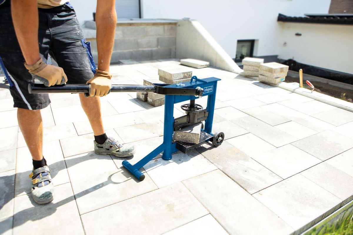 Steinknacker HSC130 von Scheppach im Einsatz. Ein Arbeiter bedient die Maschine auf einer Terrasse. Im Hintergrund sind Pflastersteine und Werkzeuge zu sehen.