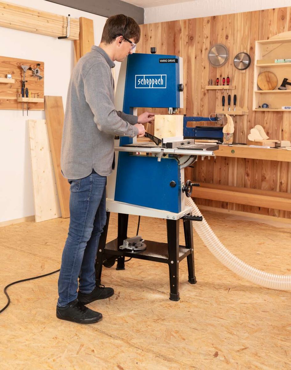 Ein Mann bedient eine Bandsäge BASA3 von Scheppach in einer Werkstatt. Die Maschine hat ein blaues Gehäuse mit weißem Logo. Im Hintergrund sind Werkzeuge und Holzregale zu sehen.