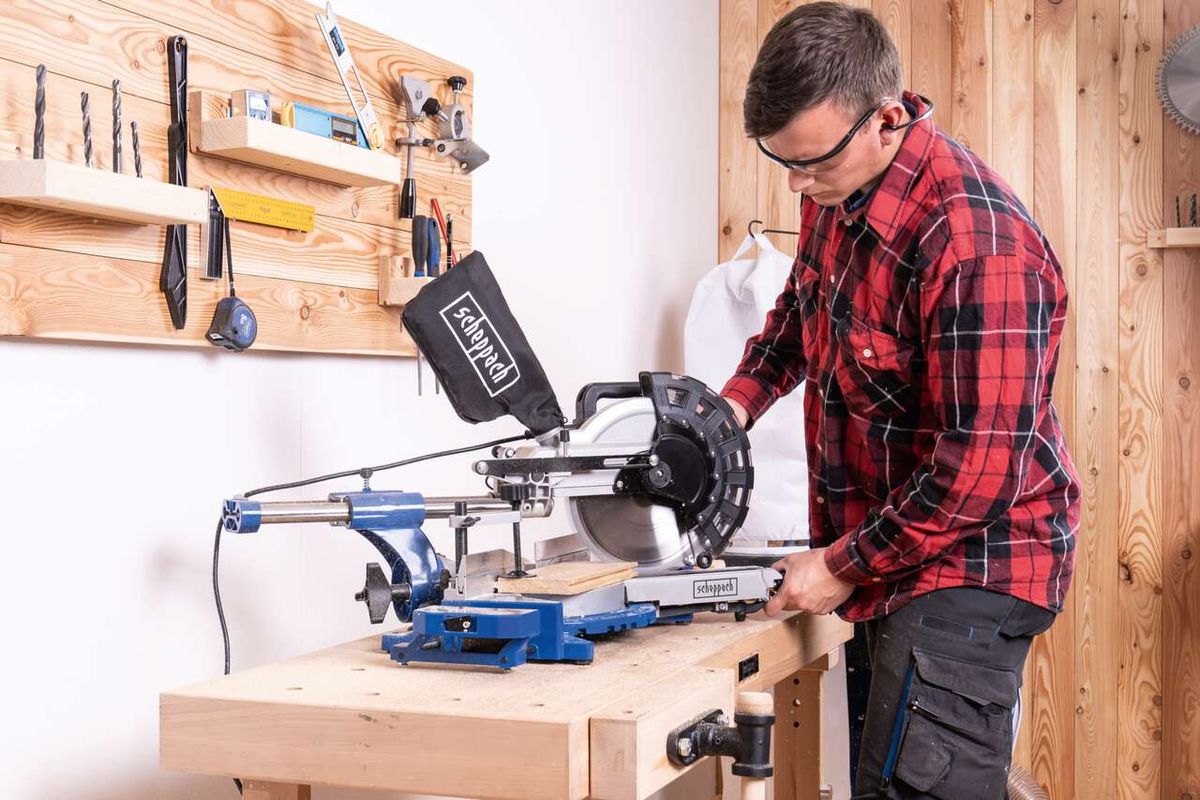 Ein Mann bedient eine Kappzugsäge HM254 von Scheppach in einer Werkstatt. Die Säge steht auf einer Werkbank und ist mit einem Holzbrett im Einsatz. Im Hintergrund sind Werkzeuge an der Wand befestigt.