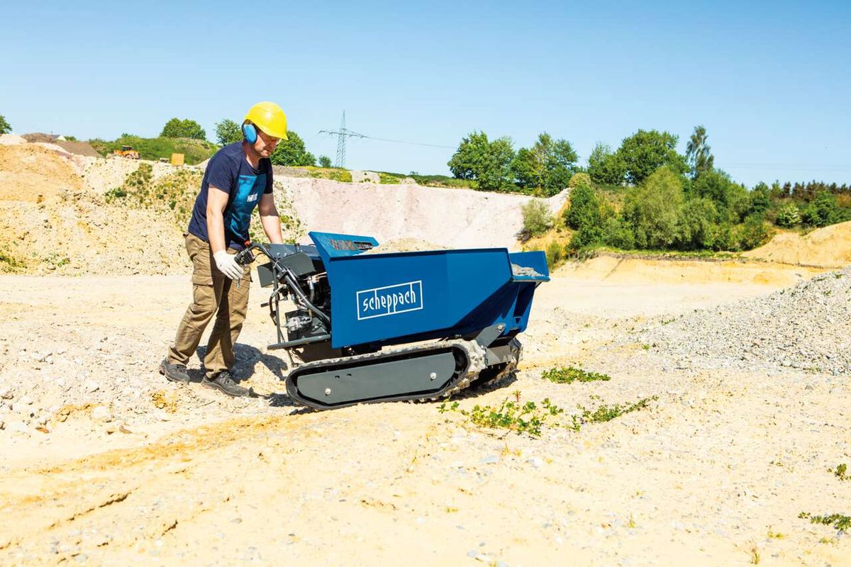 Kettendumper DP5000 Scheppach im Einsatz auf einer Baustelle. Ein Arbeiter in Schutzausrüstung bedient das Gerät auf einem unebenen Gelände. Scheppach-Logo sichtbar auf dem Behälter.
