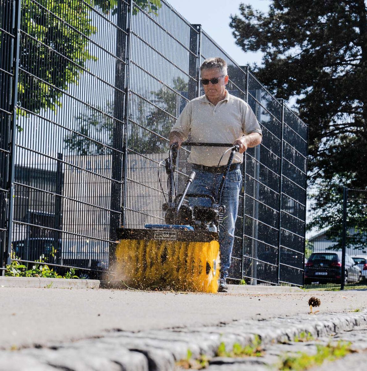Ein Mann bedient die Kehrmaschine SC2400p von Scheppach auf einem Gehweg. Die Maschine ist in Betrieb und wirft Schmutz auf. Im Hintergrund ist ein Zaun und ein Auto zu sehen.