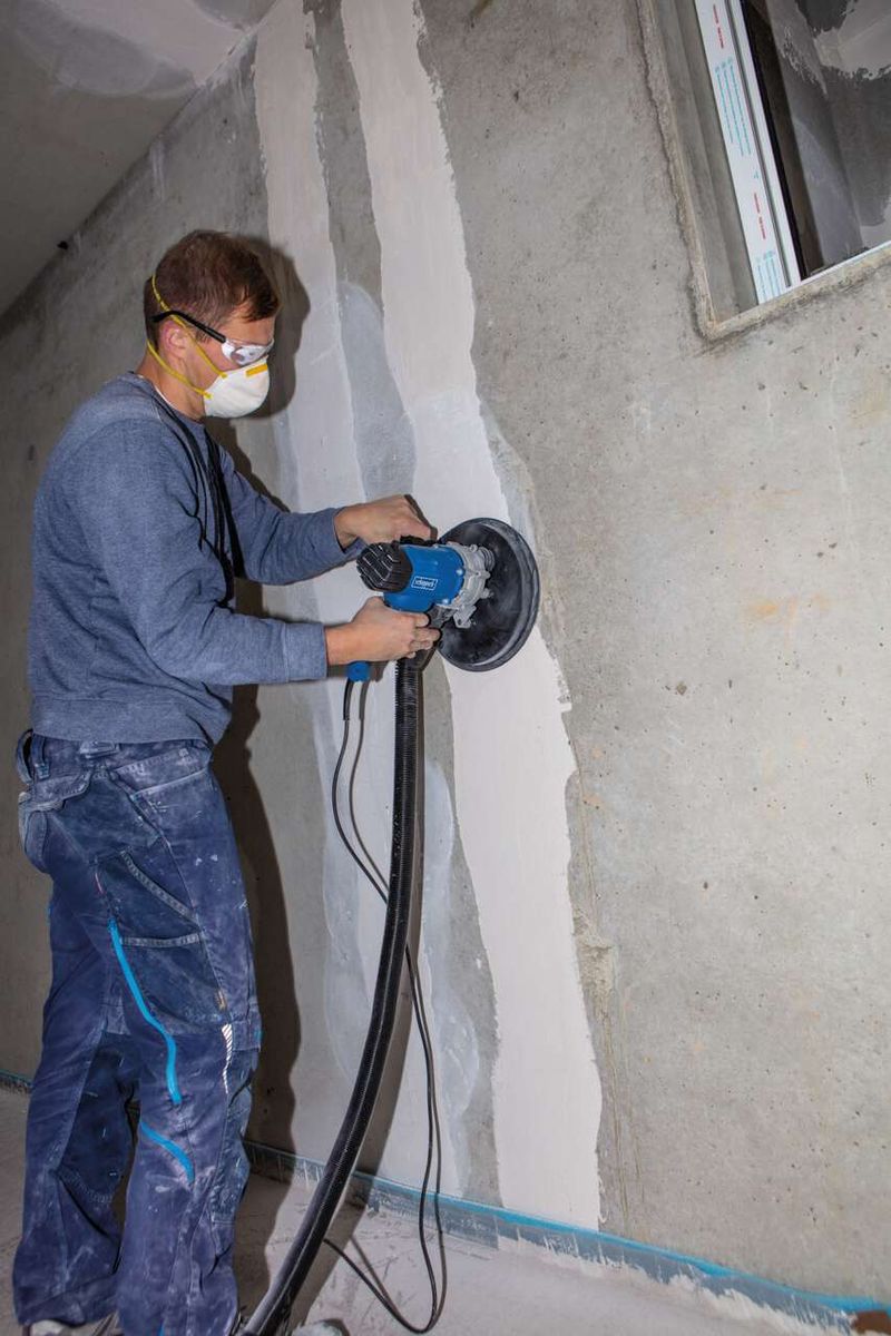 Ein Arbeiter nutzt den Wand- und Deckenschleifer DS200 von Scheppach an einer Wand. Der Schleifer hat einen Durchmesser von 215 mm und eine Leistung von 1200 W. Der Arbeiter trägt Schutzausrüstung.