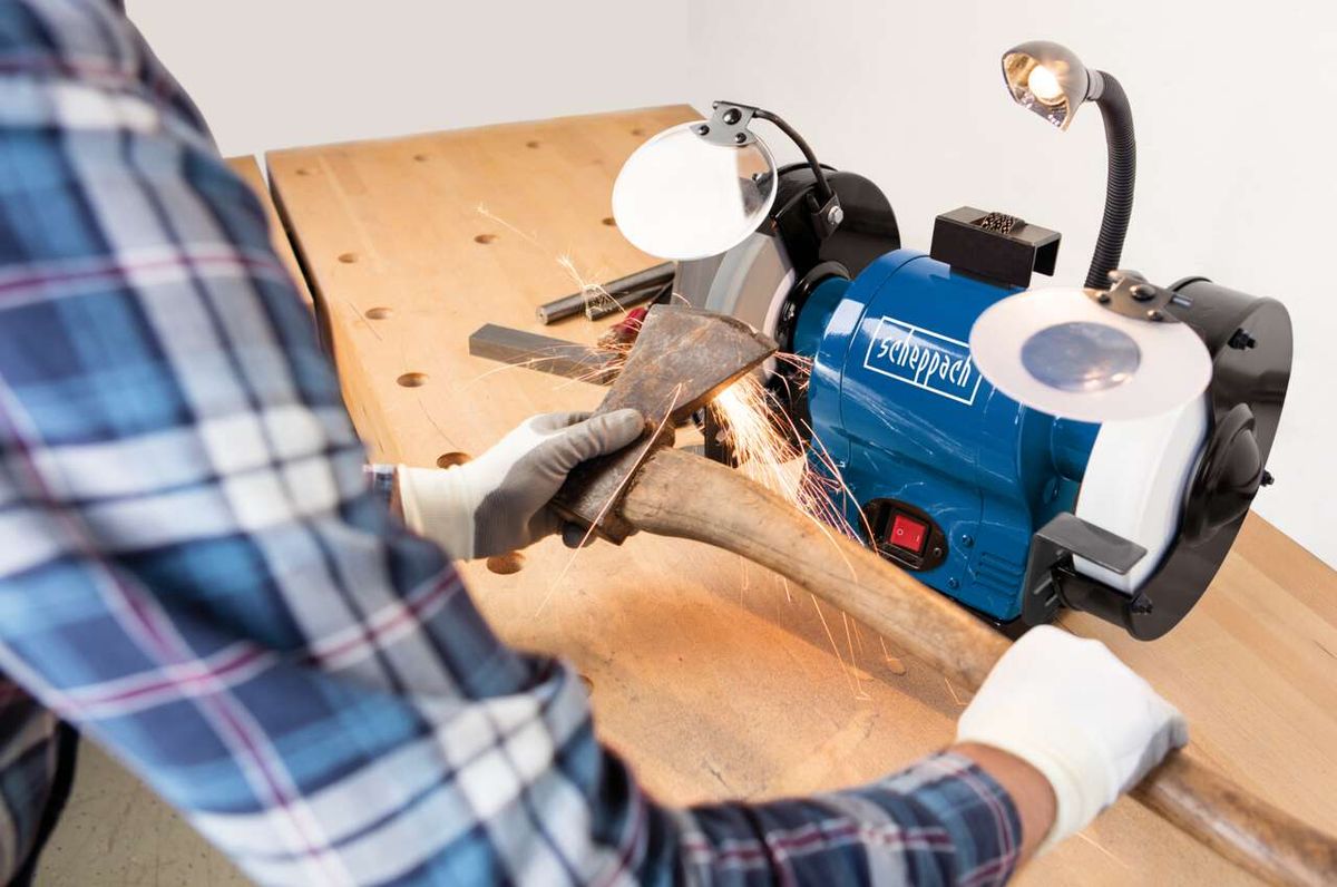 Ein Mann verwendet den Scheppach Doppelschleifer BG200AL zum Schleifen eines Metallteils. Der Schleifer ist auf einem Holztisch montiert und mit einer Lampe beleuchtet. Der Benutzer trägt Handschuhe und eine karierte Schürze.