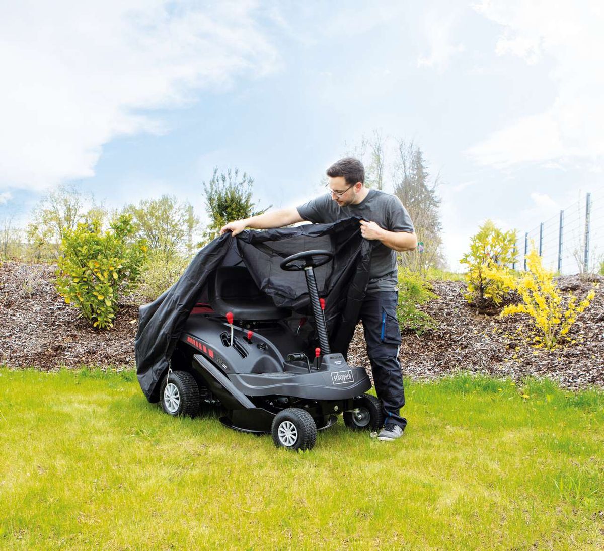 Ein Mann deckt den Scheppach Aufsitzrasenmäher MR224-61 ab. Der Rasenmäher steht auf einem Rasenstück mit jungen Pflanzen im Hintergrund.