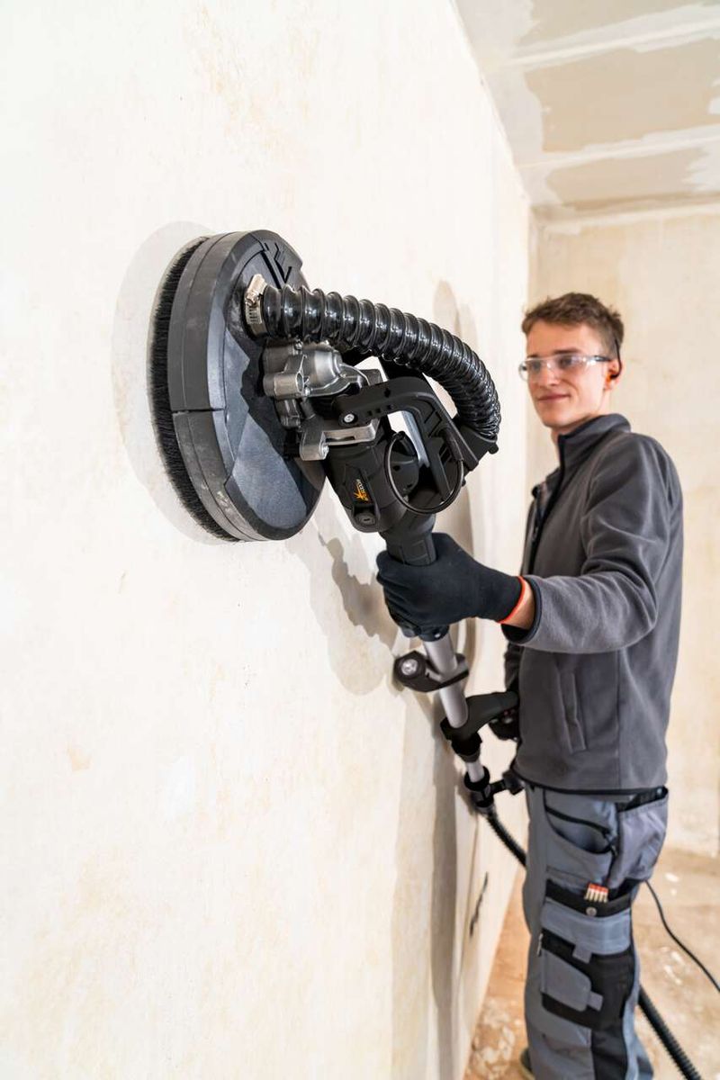 Ein Mann verwendet den Scheppach Wand- und Deckenschleifer DWS90X Woodster an einer Wand. Der Schleifer hat eine Teleskopfunktion und eine LED-Leuchte.