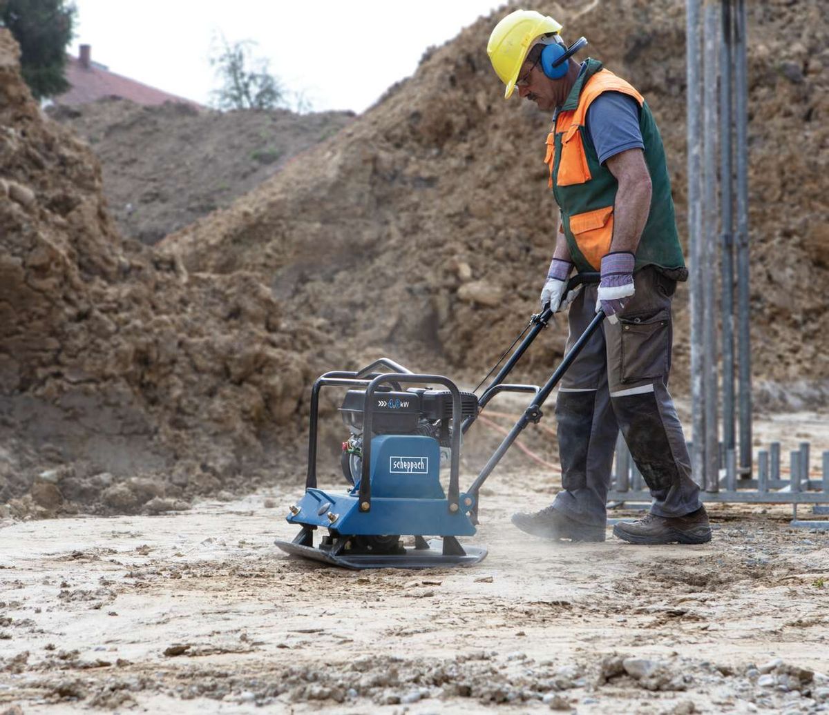 Ein Arbeiter bedient eine Scheppach Rüttelplatte HP1800S auf einer Baustelle. Die Maschine ist blau und hat einen orangefarbenen Sicherheitsvest.