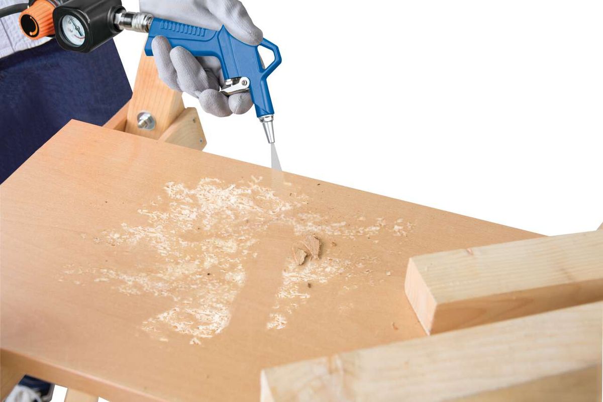 Ein Scheppach Kompressor wird verwendet, um Holz zu bearbeiten. Der Kompressor wird von einer Hand gehalten und sprüht Luft auf ein Holzbrett, das auf einer Werkbank liegt. Holzspäne sind sichtbar.