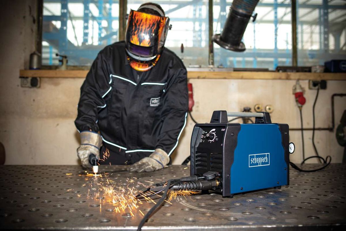 Ein Arbeiter verwendet den Plasmaschneider PLC40 von Scheppach zum Schneiden von Metall. Der Arbeiter trägt eine Schutzausrüstung, einschließlich einer Schweißerhaube und Handschuhen. Der Plasmaschneider ist auf einem Metalltisch platziert.