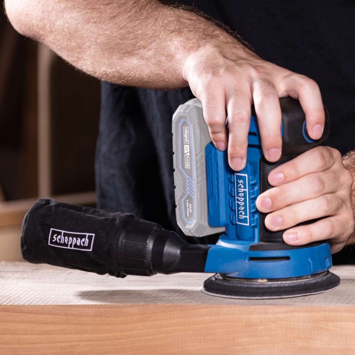 The Scheppach cordless orbital sander C-OBS125-X with 20V battery and 2.0 Ah capacity. The handle is covered with a black sleeve.