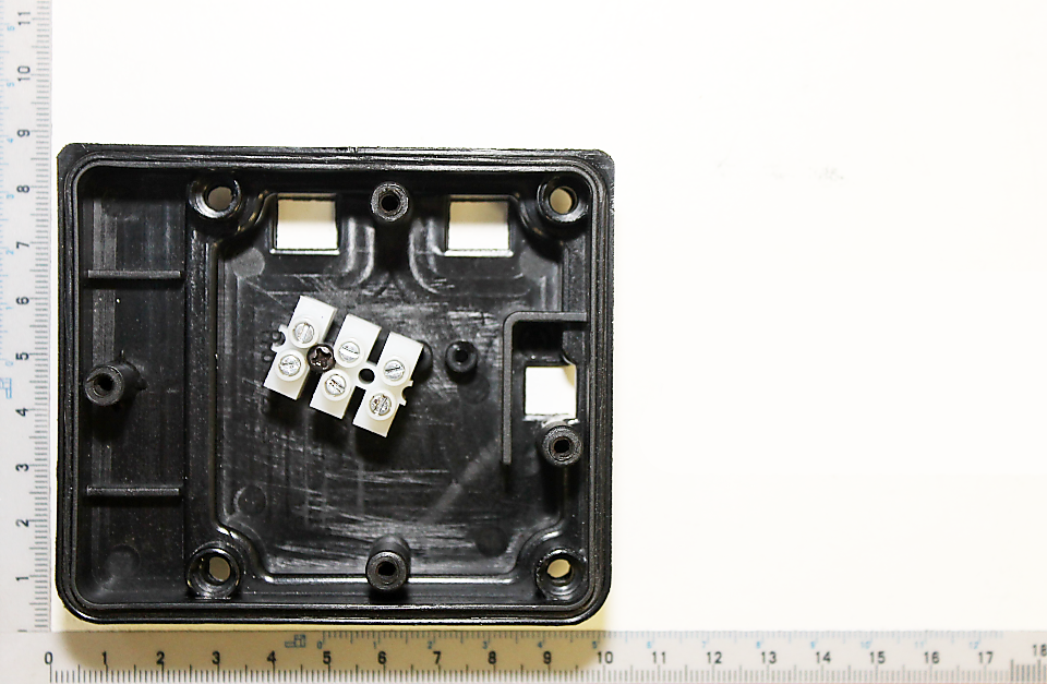 Kabelbox Abdeckung von Scheppach, Ansicht von oben. Die Abdeckung ist rechteckig mit vier Befestigungslöchern an den Ecken und einem zentralen Bereich mit elektronischen Komponenten. Die Maße betragen etwa 9 cm x 7 cm.