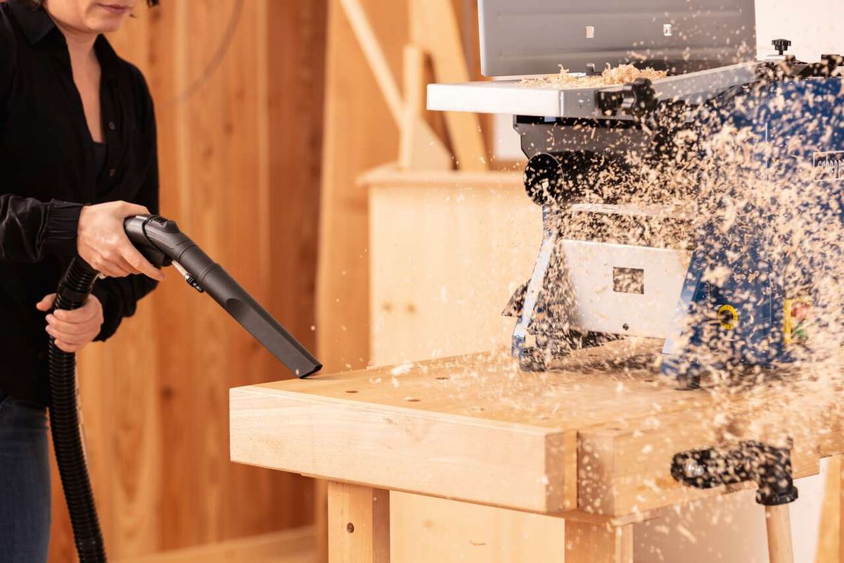 Ein Scheppach SprayVac20-Sauger saugt Holzspäne von einer Werkbank. Im Hintergrund ist eine Kreissäge zu sehen.