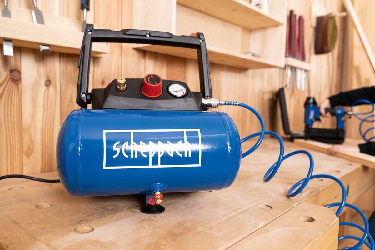 Scheppach Kompressor HC06 auf einer Werkbank. Der Kompressor hat einen blauen Tank und einen schwarzen Kopf mit einem roten Druckregler. Ein blauer Schlauch ist angeschlossen.