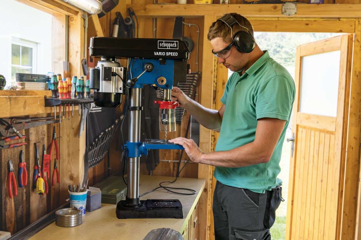 A man operates a Scheppach bench pillar drill DP19Vario in a workshop. The man is wearing safety goggles and ear protection. The workbench is equipped with tools and accessories.