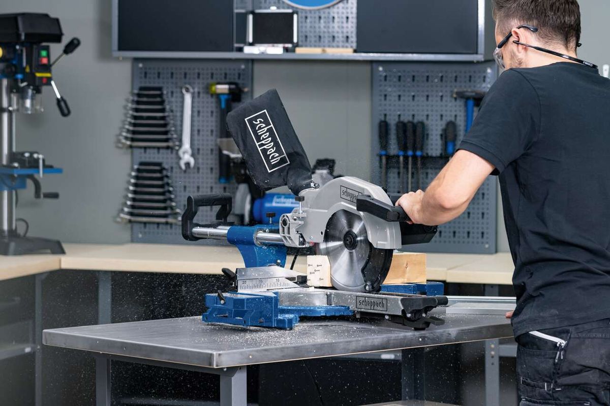 Ein Mann bedient eine Scheppach-Kappzugsäge HM110MP in einer Werkstatt. Die Säge steht auf einer Metallplatte und hat eine schwarze Schutzhaube. Im Hintergrund sind Werkzeuge an der Wand aufgehängt.
