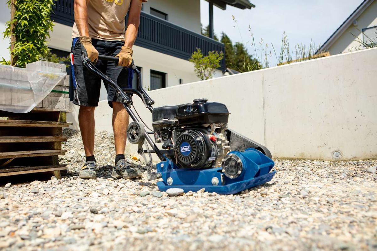 Rüttelplatte HP1900S von Scheppach im Einsatz, Person bedient die Maschine auf einem Kiesboden. Die Maschine hat einen schwarzen Motor mit blauem Gehäuse.