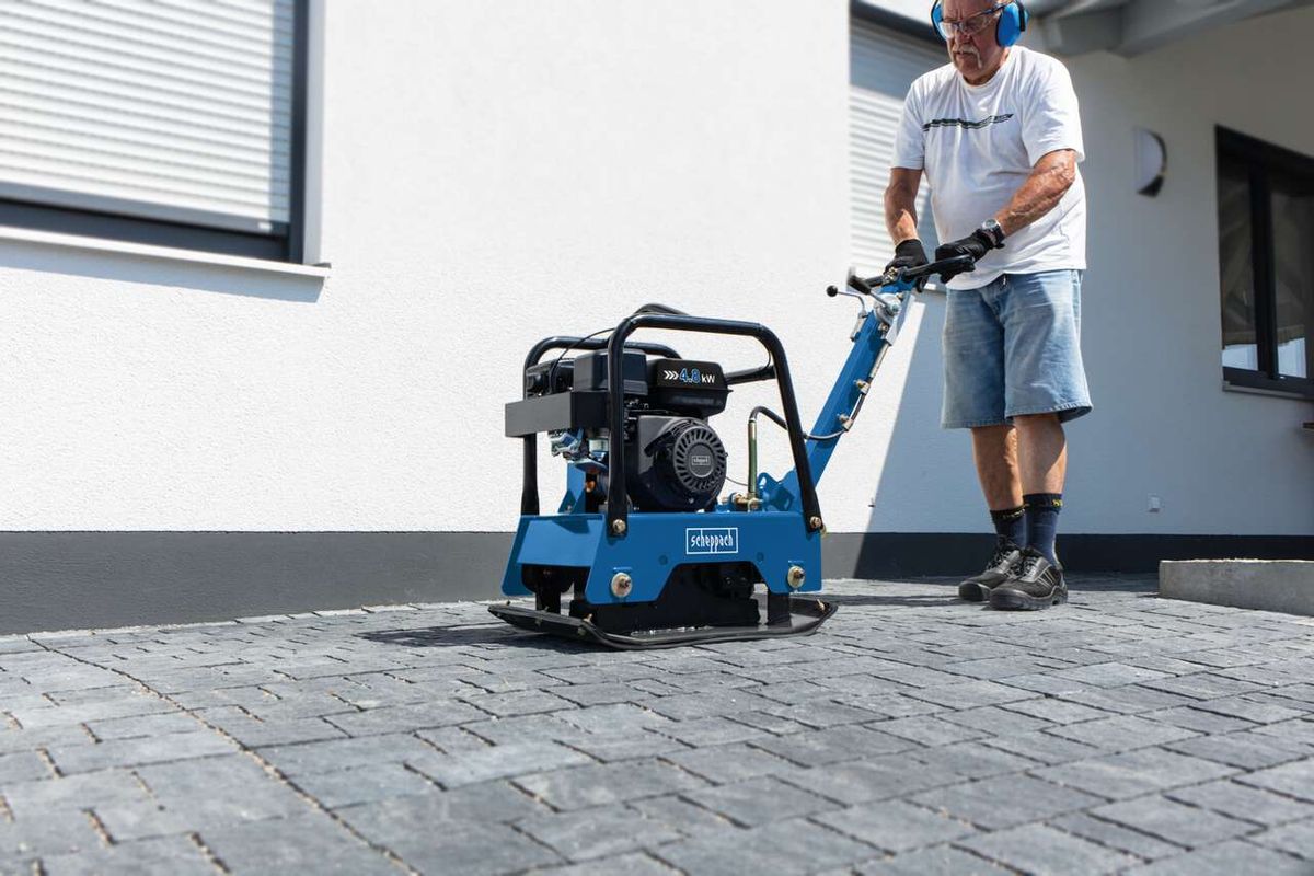 Ein Mann bedient eine Scheppach Rüttelplatte HP2500S vor einem Gebäude. Die Maschine hat einen schwarzen Motor und eine blaue Basisplatte.