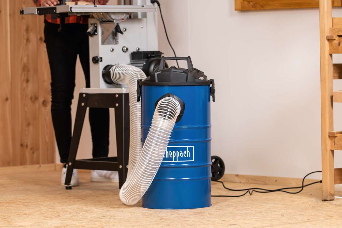 Scheppach DC100 dust extractor with blue drum and hose connection. Person standing next to it operating a power tool.