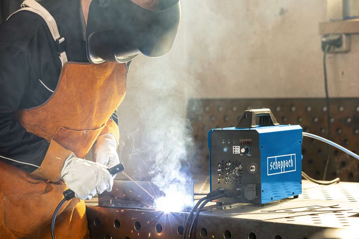 Ein Arbeiter schweißt mit dem 3in1 Kombi-Schweißgerät WSE4000-CUT von Scheppach. Der Schweißer trägt Schutzausrüstung und arbeitet an einem Metallgitter. Das Gerät steht auf dem Tisch und erzeugt Rauch.