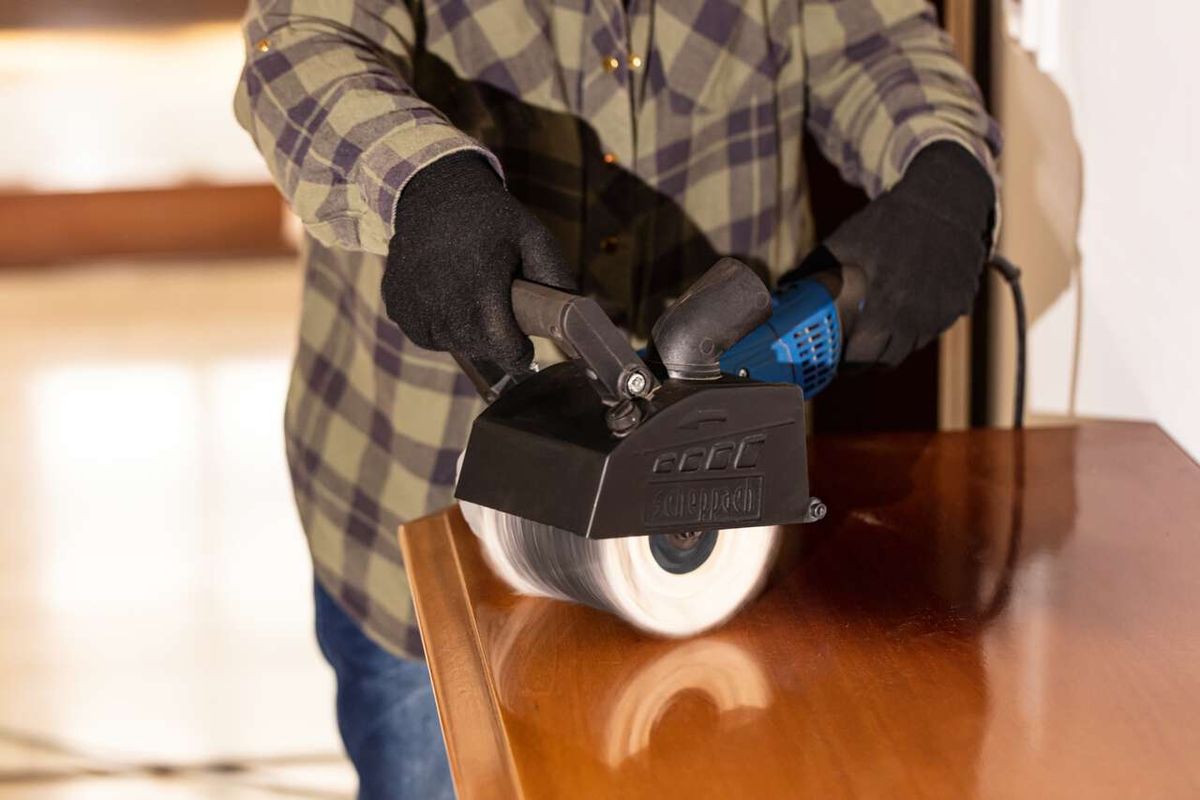 Ein Mann hält eine Scheppach Schleifmaschine mit einer Baumwollbürstenaufsatz. Die Maschine ist in Betrieb und wird auf einer Holzfläche verwendet.