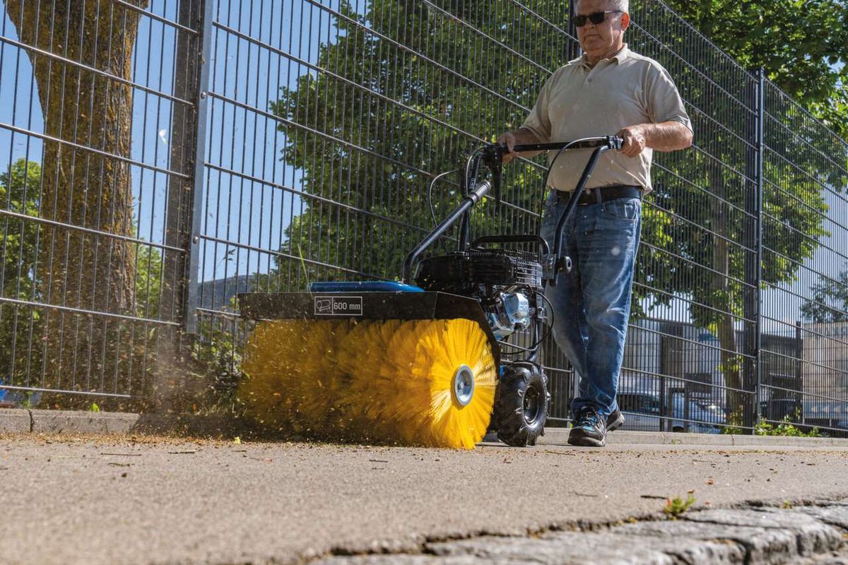 Ein Mann bedient eine Kehrmaschine SC2400p von Scheppach auf einem Gehweg. Die Maschine hat eine gelbe Bürste und wirft Laub zur Seite.